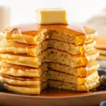 A stack of golden Kodiak Cakes pancake topped with butter, fresh blueberries, and maple syrup, served on a rustic wooden plate.